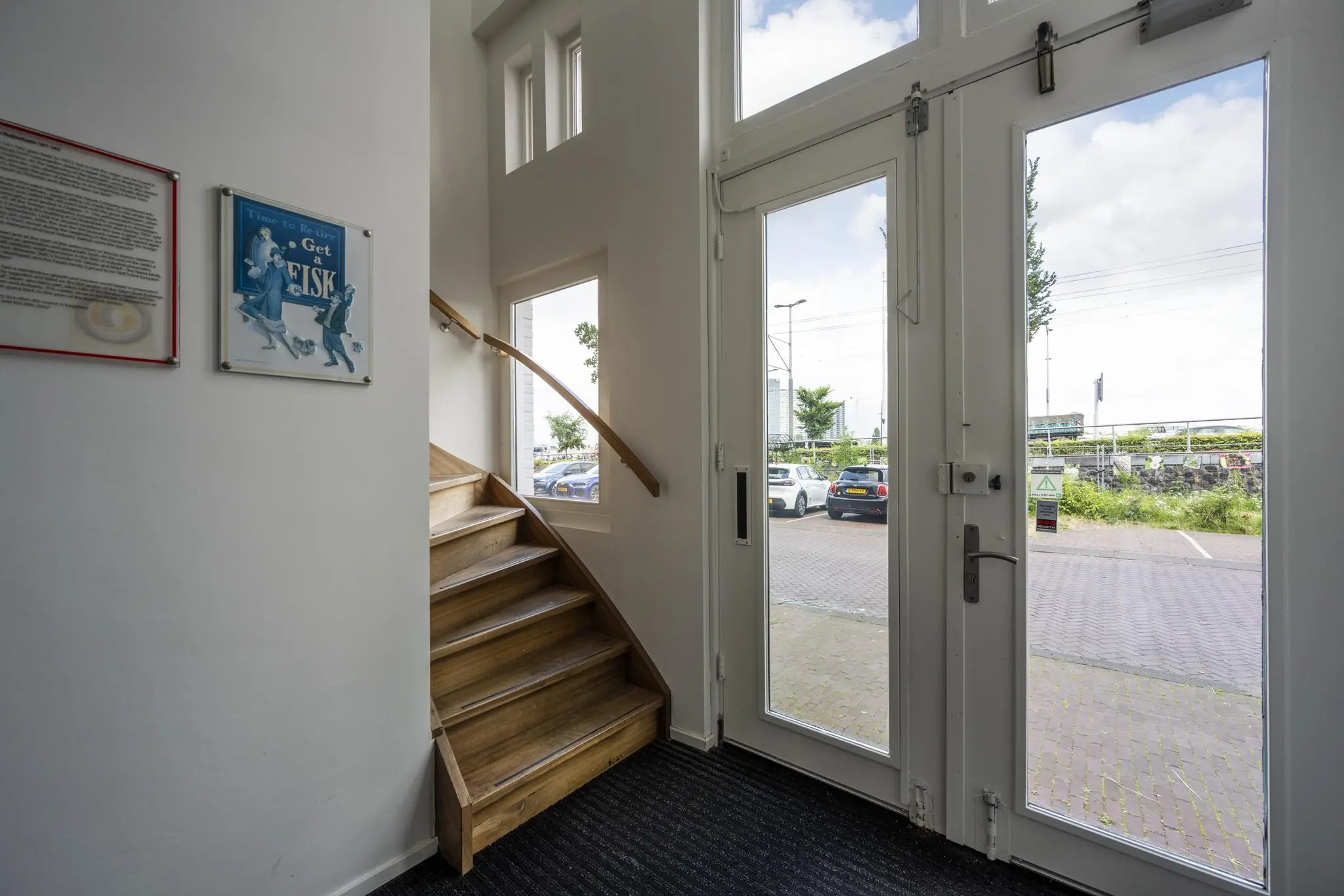 Entreehal met houten trap, glazen toegangsdeur en uitzicht op parkeerplaats aan de De Ruijterkade.